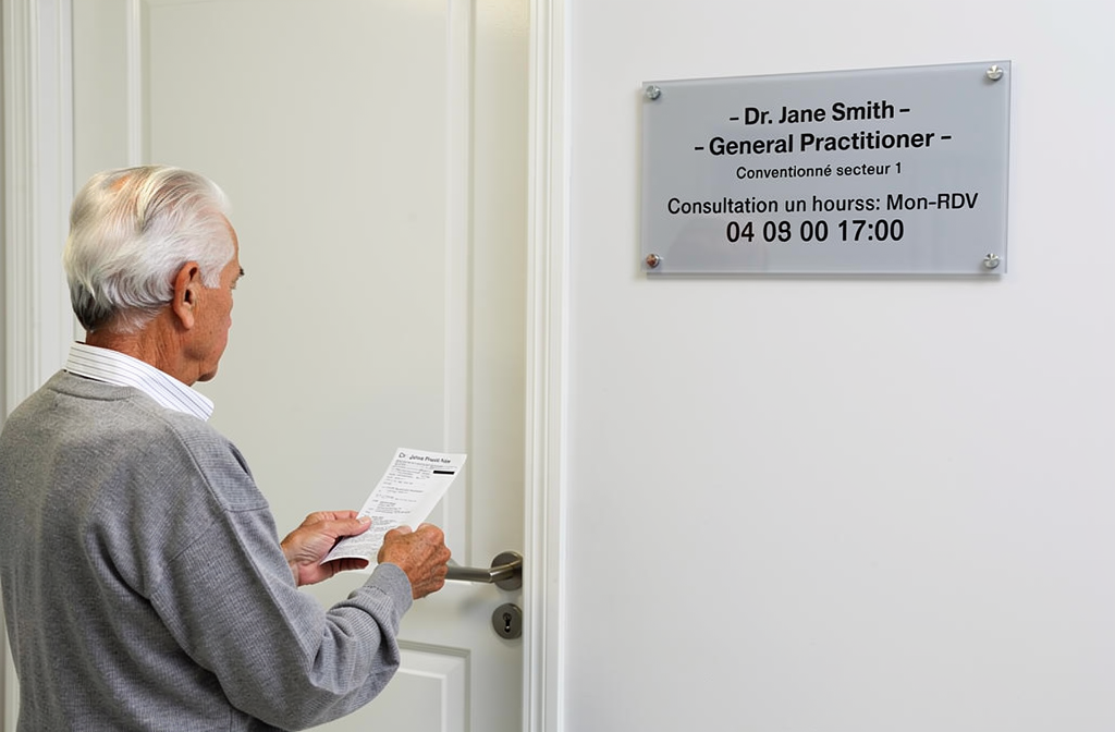 Homme âgé tenant une ordonnance papier devant une porte de cabinet médical, plaque d’information sur le mur. integration keyword: comment l'ordonnance papier renforce-t-elle l'observance thérapeutique chez les patients âgés