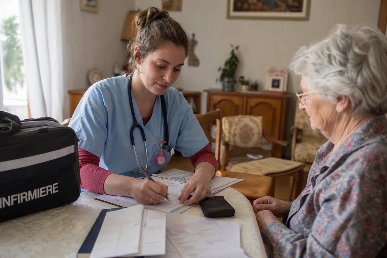 Infirmière libérale à domicile rédigeant des factures et une feuille de soins pour une patiente âgée.